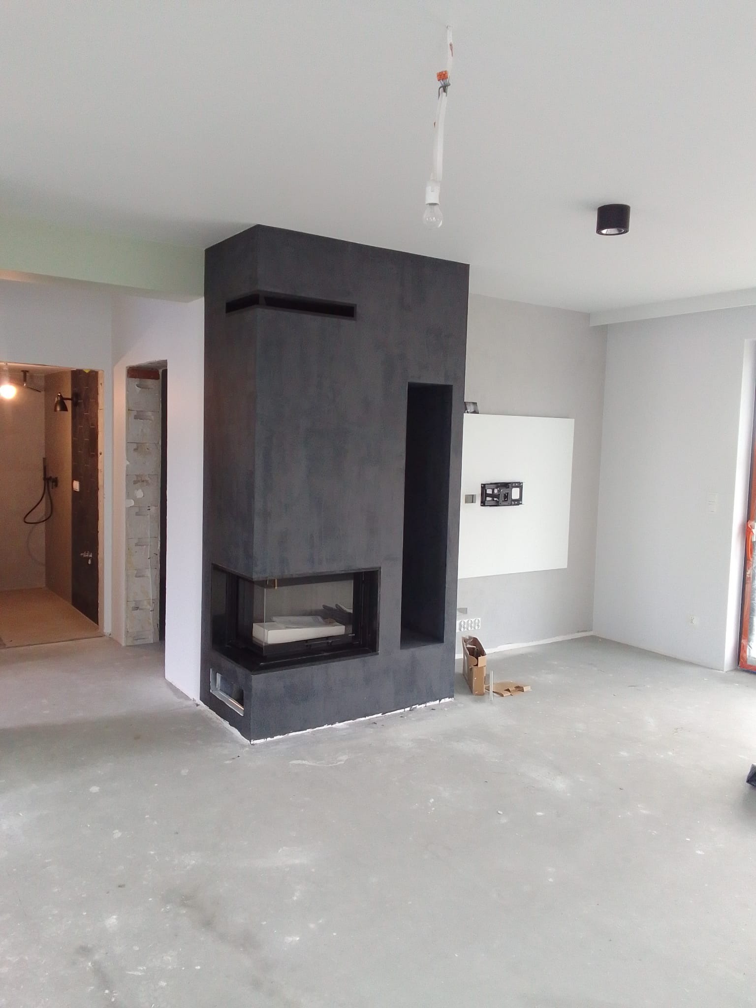 Nowoczesny, surowy kominek z czarnego betonu architektonicznego w jasnym, surowym wnętrzu w trakcie wykańczania, widoczne niedokończone podłogi i instalacje elektryczne.