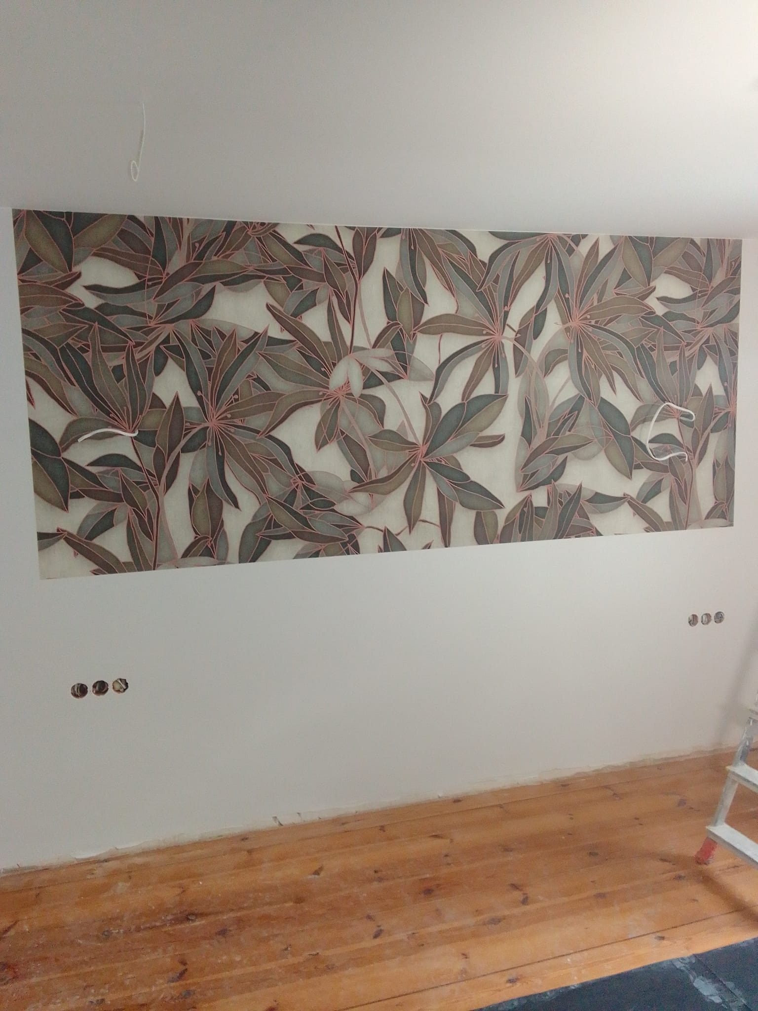 Tapeta z motywem liści na ścianie, widoczne niedokończone gniazdka elektryczne i fragment drewnianej podłogi, drabina malarska z boku