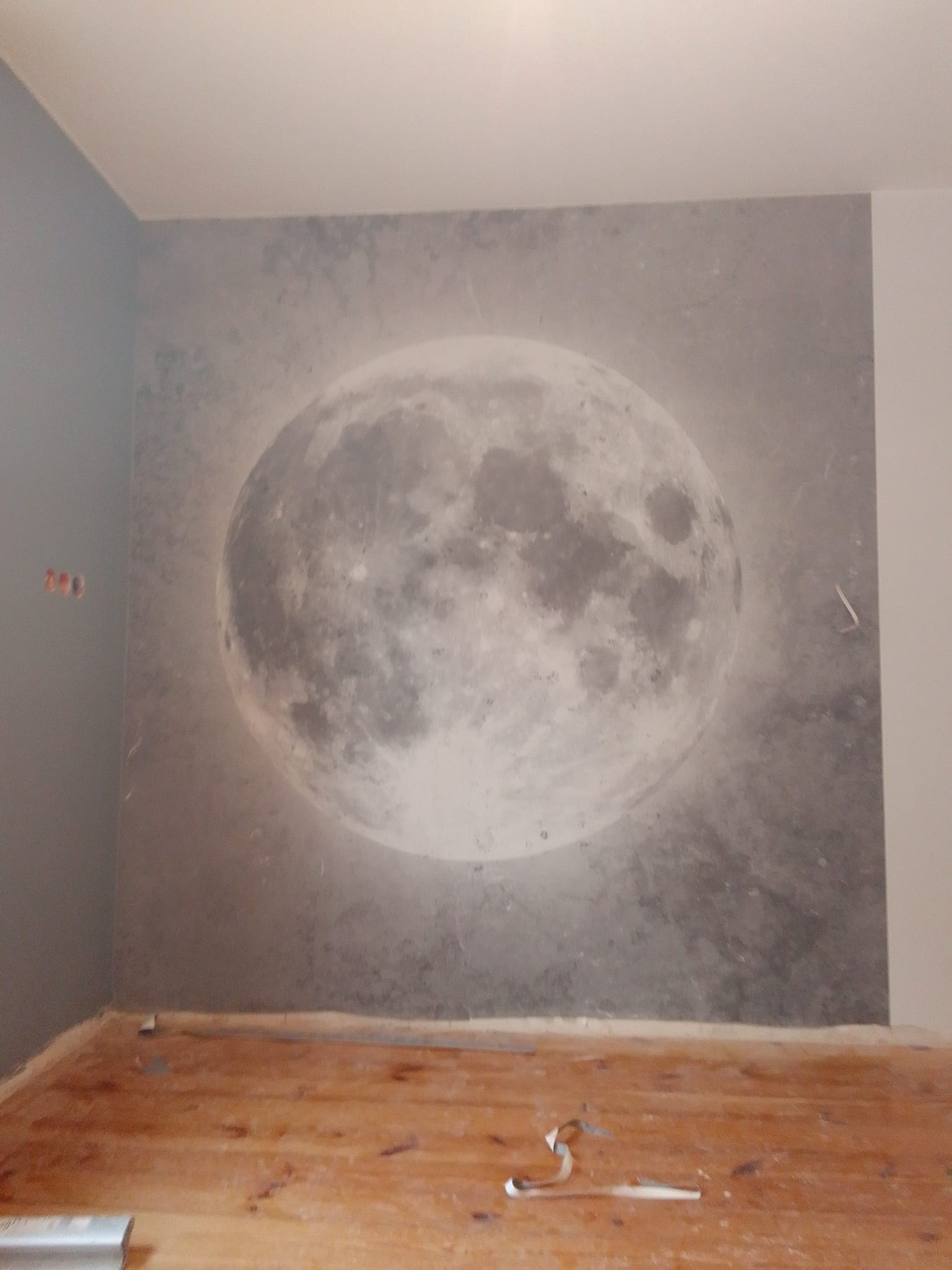 Ściana z fototapetą przedstawiającą księżyc w szarościach, świeżo położona podłoga z desek, widoczne ślady po remoncie.