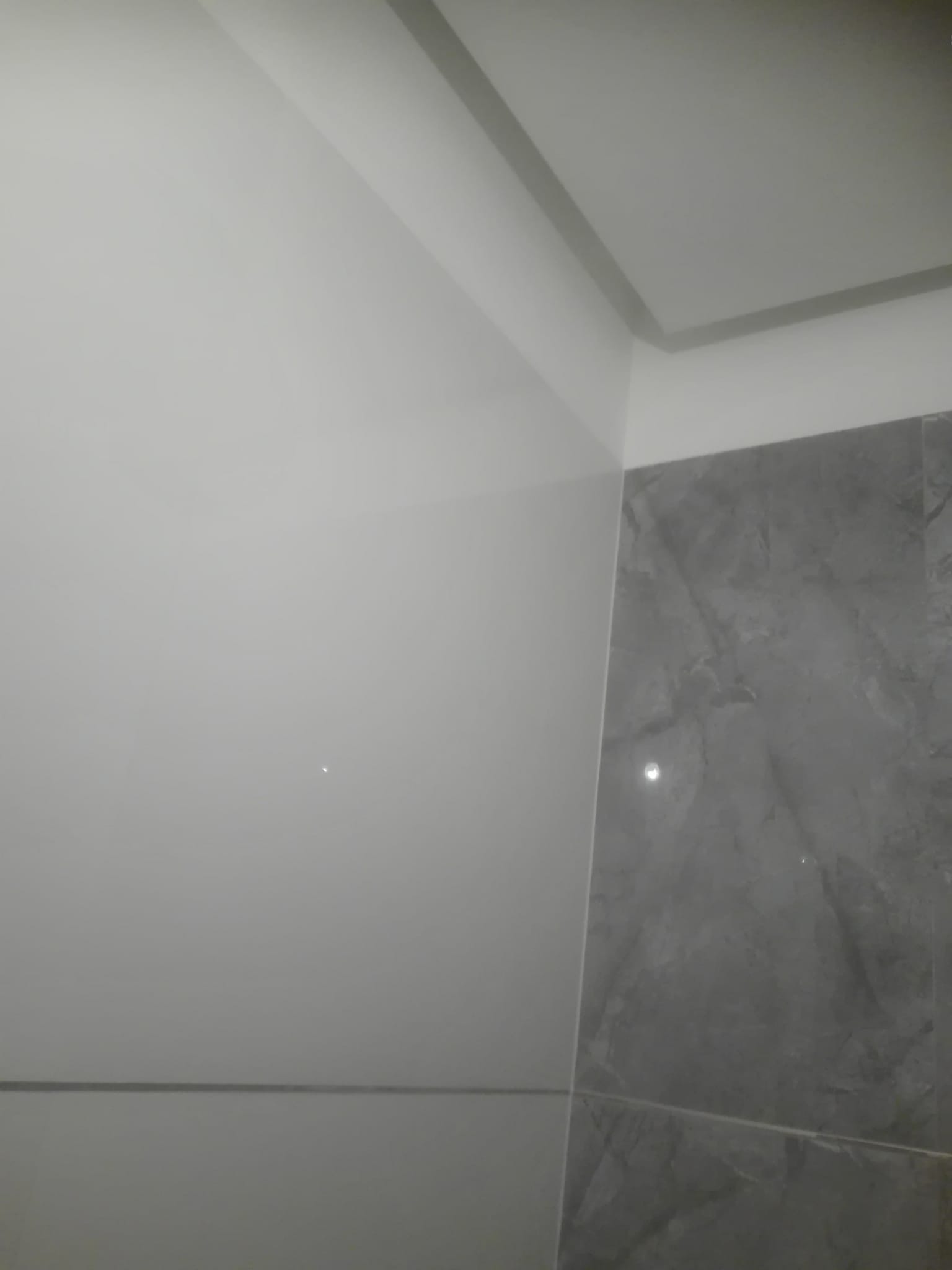Fragment ściany wykończonej jasnymi płytkami ceramicznymi i szarą płytką imitującą marmur, widoczny narożnik sufitu z dekoracyjną listwą.