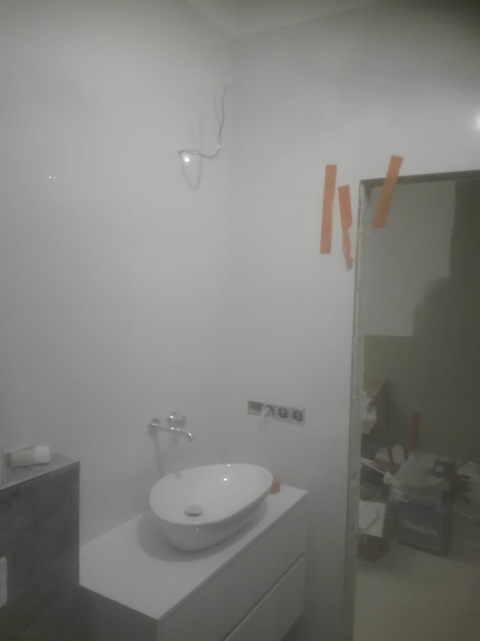 Łazienka w trakcie prac wykończeniowych: biała umywalka nablatowa na szafce, widoczne kable ze ściany, otwory na gniazdka elektryczne i taśma malarska przy framudze drzwi.