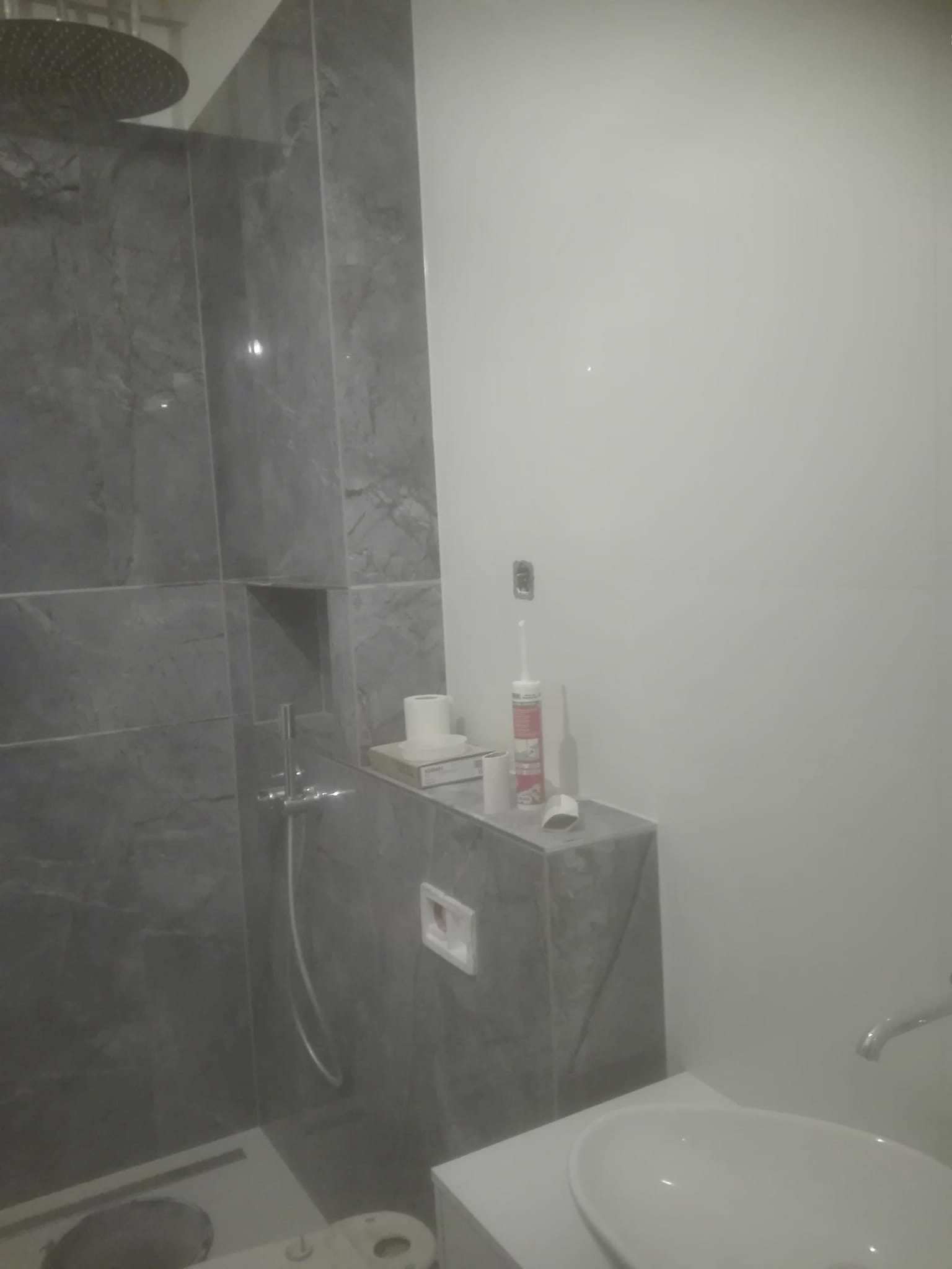 Szare płytki imitujące marmur w kabinie prysznicowej w Legnicy, widoczna bateria, odpływ liniowy i biała ściana z pojemnikiem silikonu.