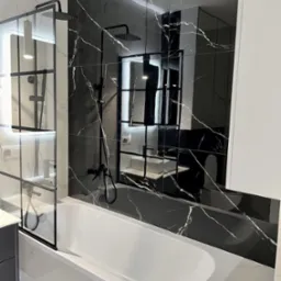 Nowoczesna łazienka z białą wanną, czarną armaturą i płytkami imitującymi marmur w odcieniach czerni i bieli, kabina prysznicowa ze szklaną ścianką z czarnymi profilami.