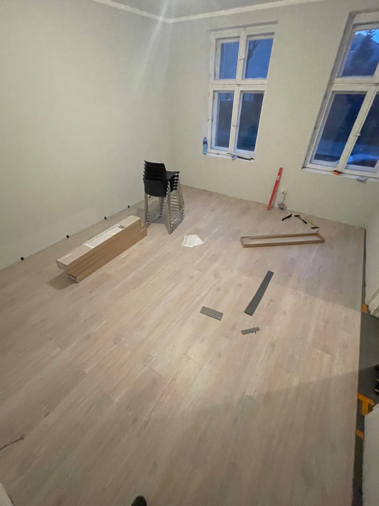 Jasne panele podłogowe w trakcie układania w pokoju z dwoma oknami, widoczne narzędzia, listwy przypodłogowe, paczka paneli i krzesła w tle.