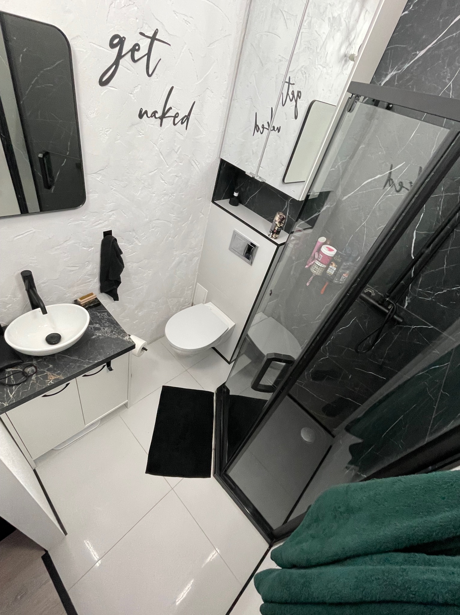 Nowoczesna łazienka z czarnymi akcentami: kabina prysznicowa z marmurowym wzorem, umywalka na szafce z blatem imitującym marmur, białe płytki podłogowe, dekoracyjne napisy na ścianach.