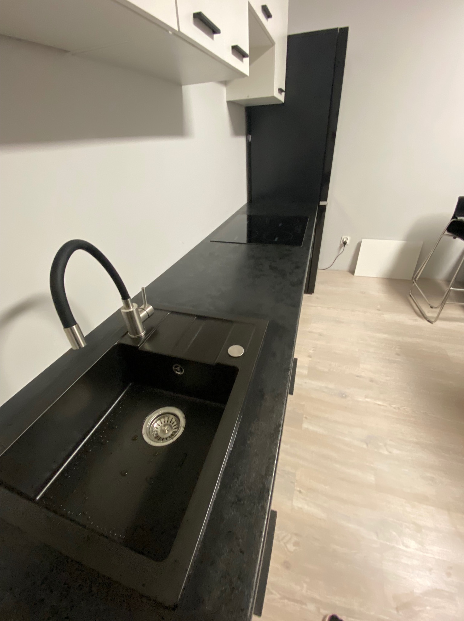 Czarny, granitowy blat kuchenny z wbudowanym zlewem i elastyczną, czarną wylewką baterii, uzupełniony płytą indukcyjną, w nowoczesnej kuchni z białymi szafkami i jasną podłogą.