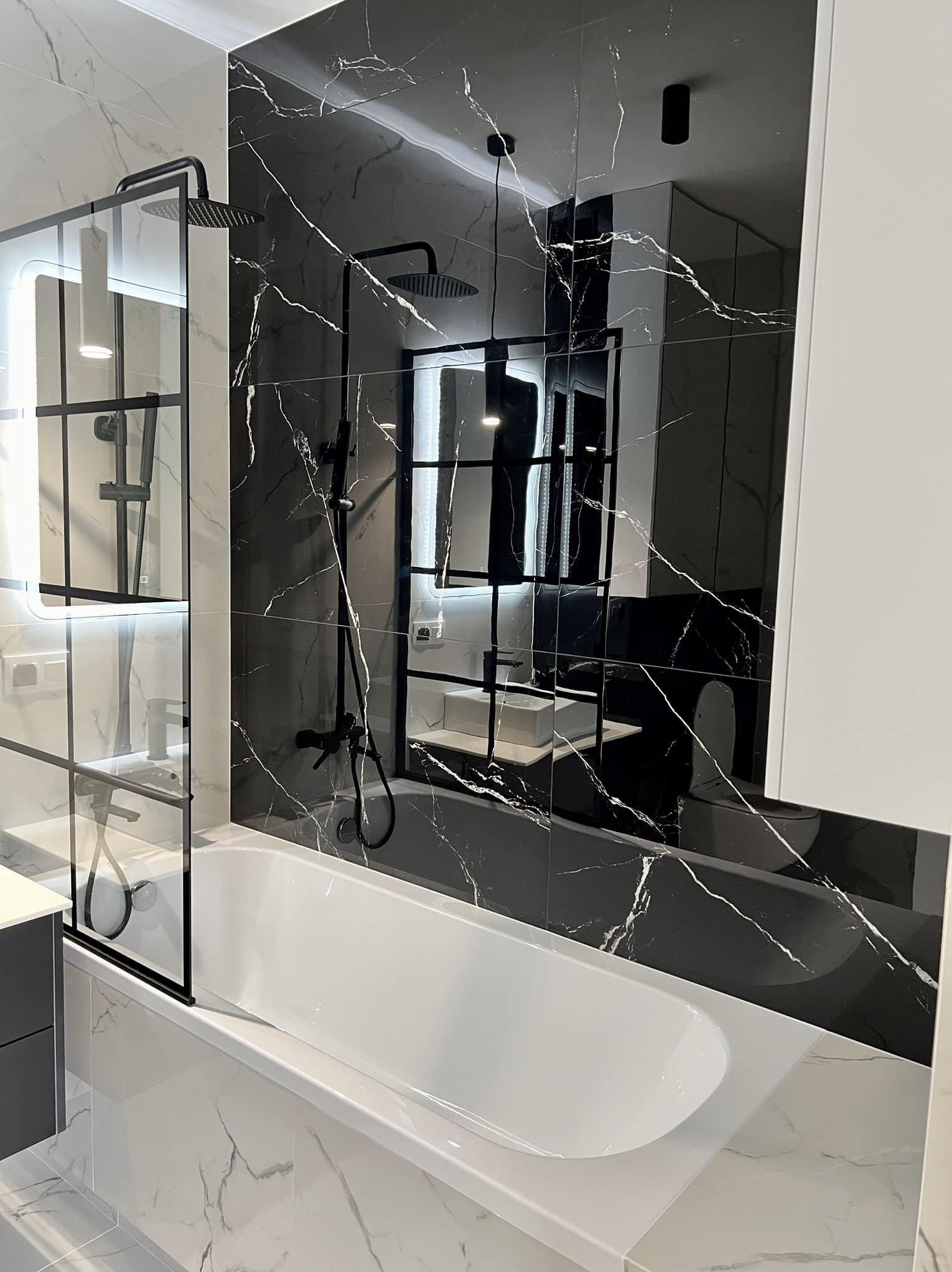 Nowoczesna łazienka z białą wanną, czarną armaturą i płytkami imitującymi marmur w odcieniach czerni i bieli, kabina prysznicowa ze szklaną ścianką z czarnymi profilami.