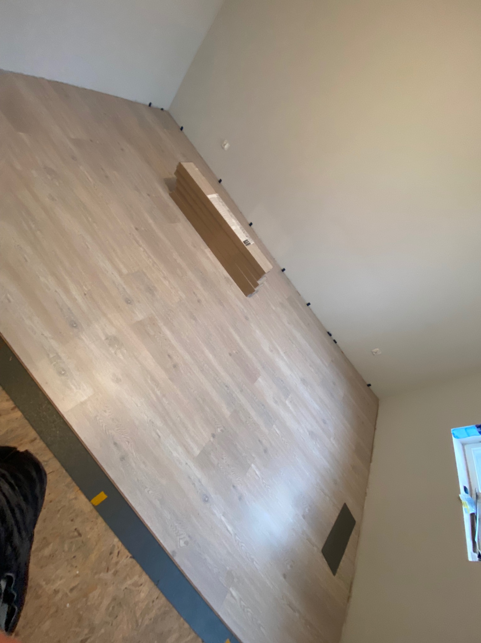 Układanie paneli podłogowych o jasnym odcieniu drewna, widoczne dystanse przy ścianie, paczka paneli oraz fragment listwy przypodłogowej w trakcie montażu.