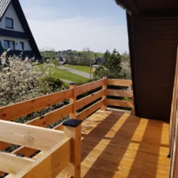 Drewniany taras z balustradą i widokiem na kwitnące drzewa oraz zabudowę jednorodzinną w oddali, słoneczny dzień.