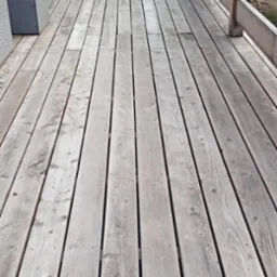 Szary taras drewniany z widoczną fakturą desek i fragmentem balustrady. W tle zieleń. Perspektywa z góry.