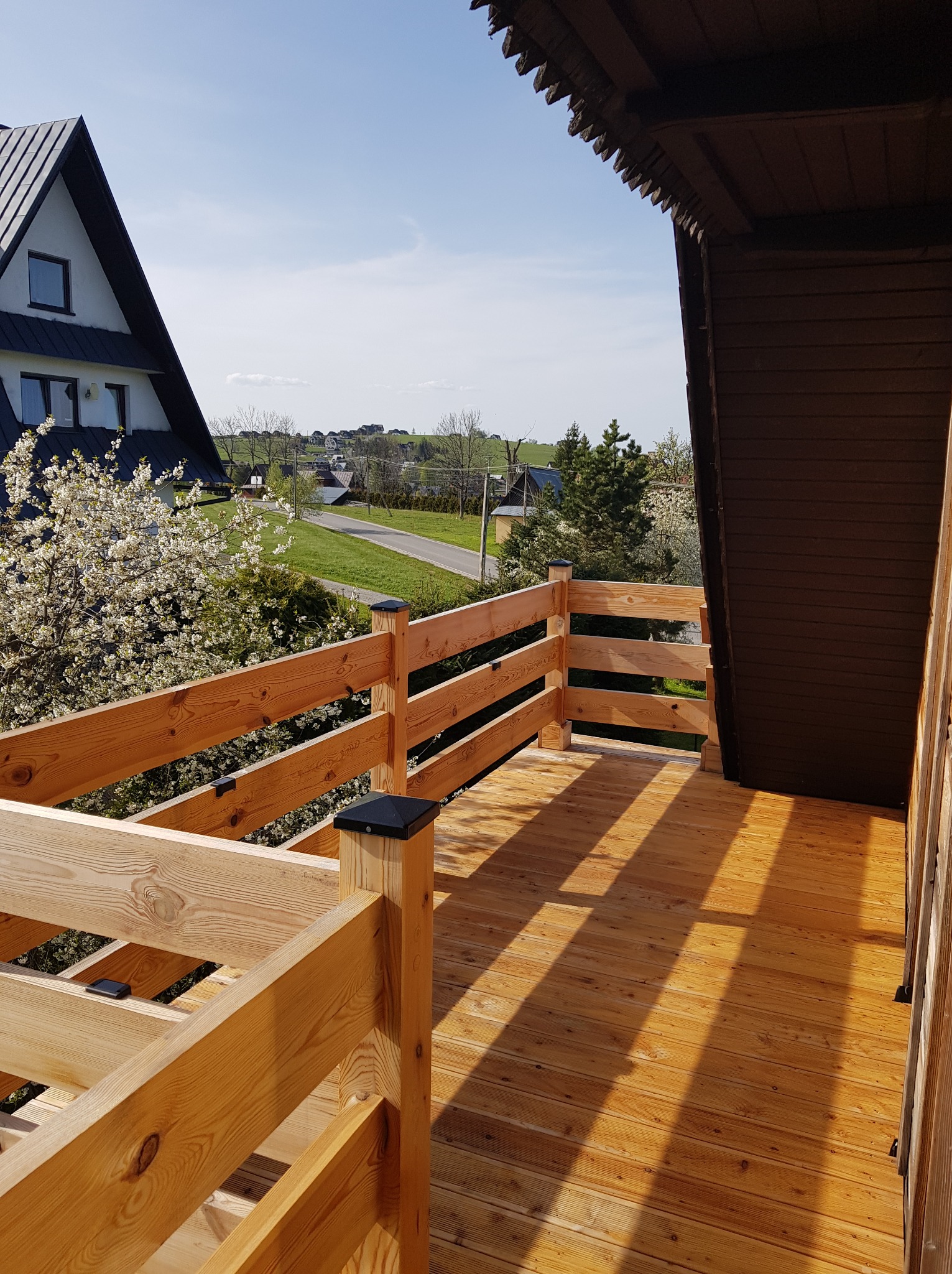 Drewniany taras z balustradą i widokiem na kwitnące drzewa oraz zabudowę jednorodzinną w oddali, słoneczny dzień.