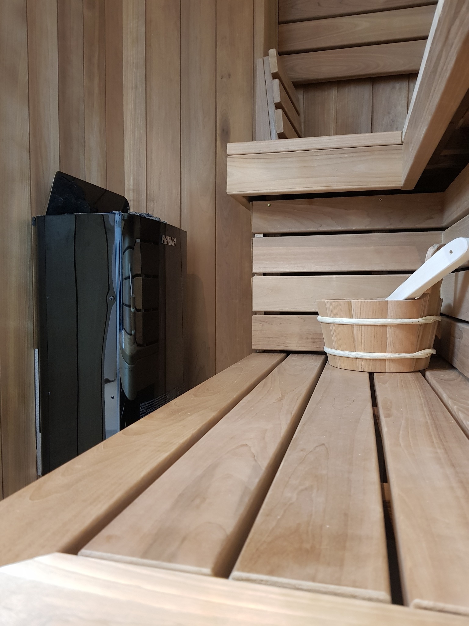 Wnętrze wykończonej sauny z drewnianymi ławkami, piecem Harvia i drewnianym wiadrem z chochlą.