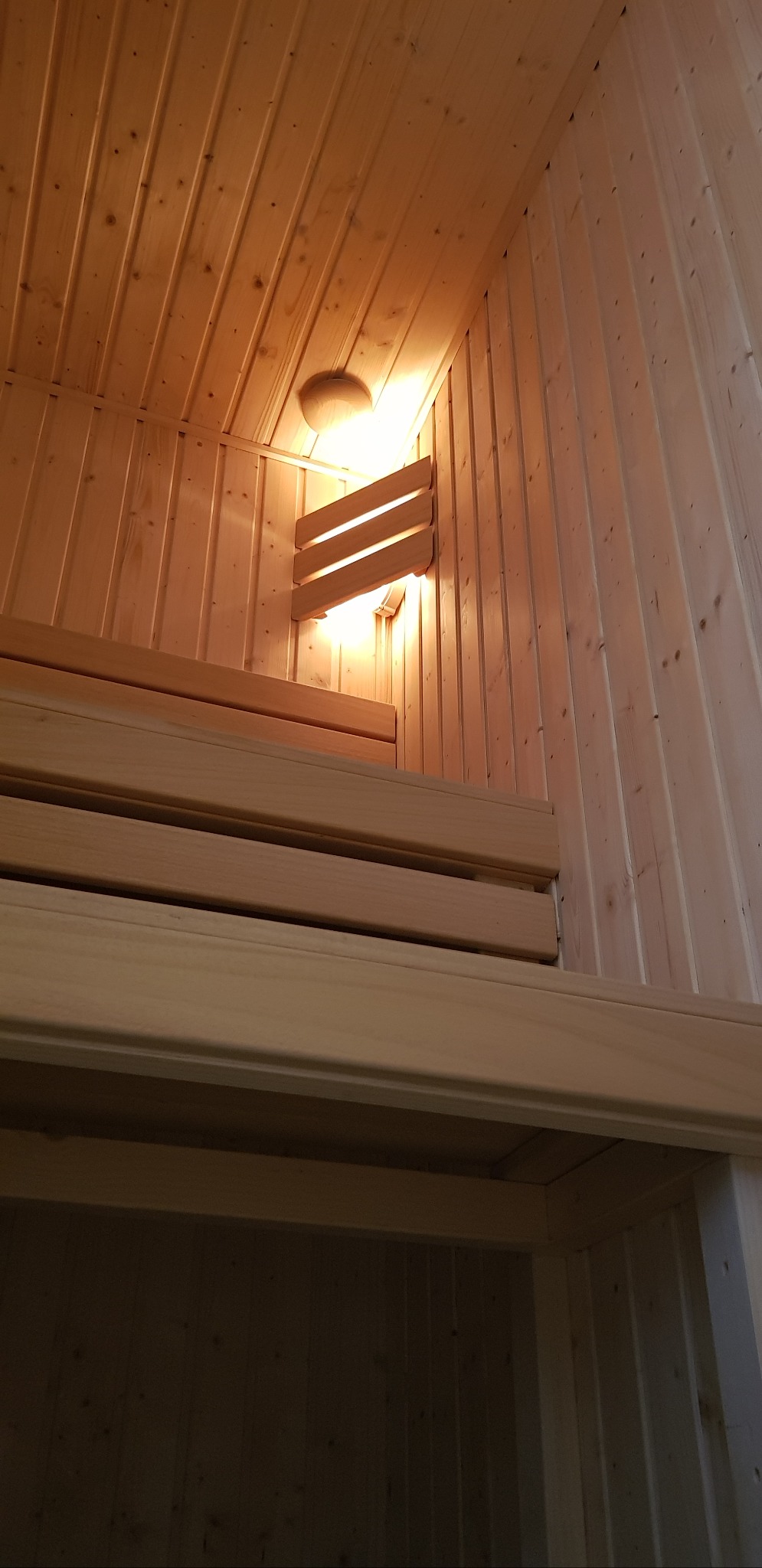 Wnętrze sauny z drewnianym wykończeniem, ławami i oświetleniem. Widok z dołu, ukazujący detale konstrukcji i ciepłe światło lampy.