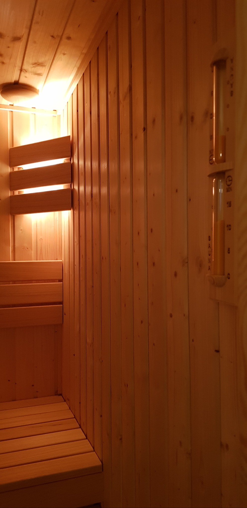 Drewniana sauna z termometrem i higrometrem. Widoczne ławy i oświetlenie. Ciepłe, pomarańczowe światło podkreśla naturalną strukturę drewna.
