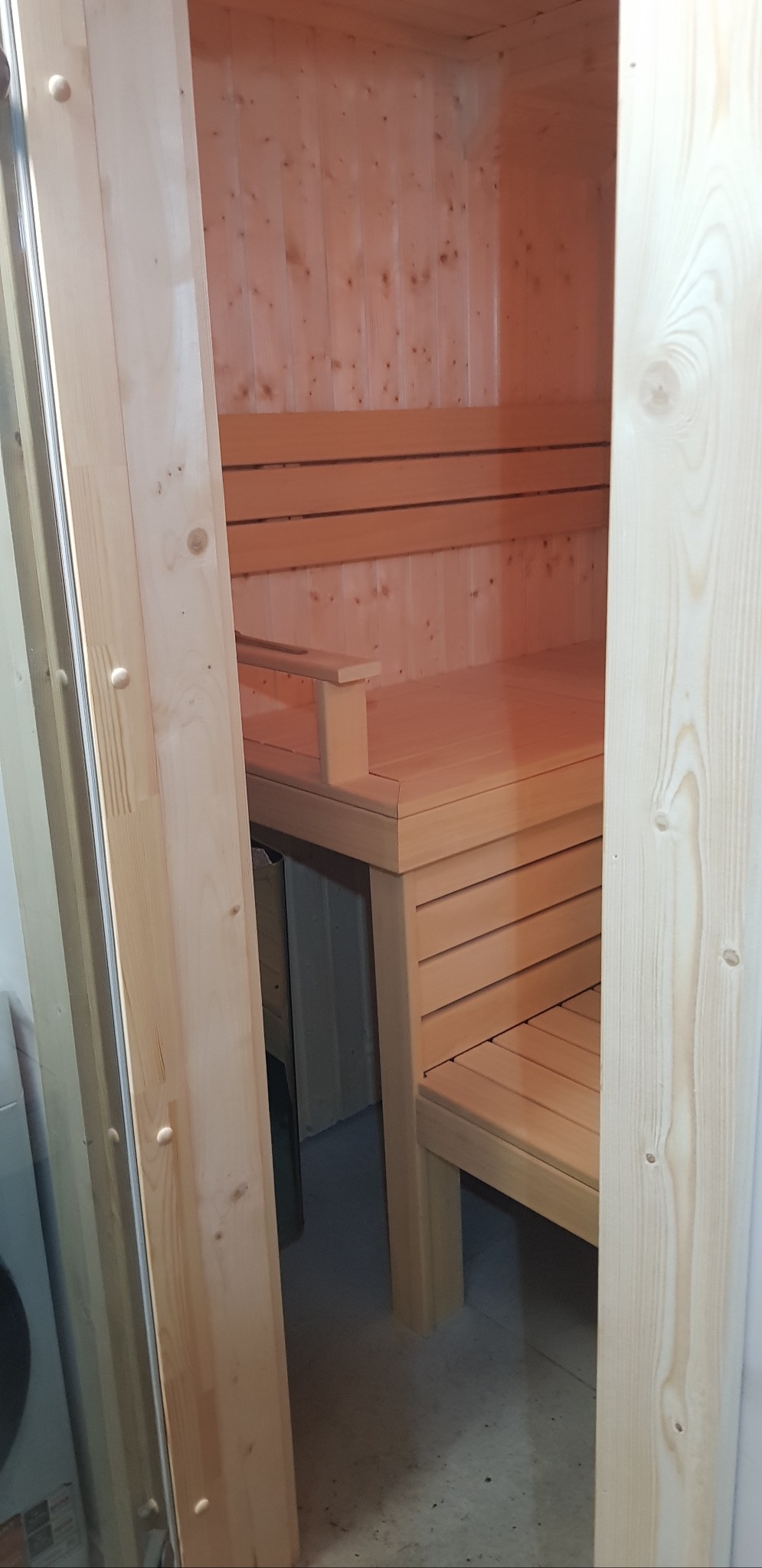 Jasne drewno i ciepłe światło wewnątrz sauny. Widoczne ławy z poręczą i fragment pieca. Drzwi sauny z widoczną strukturą drewna.