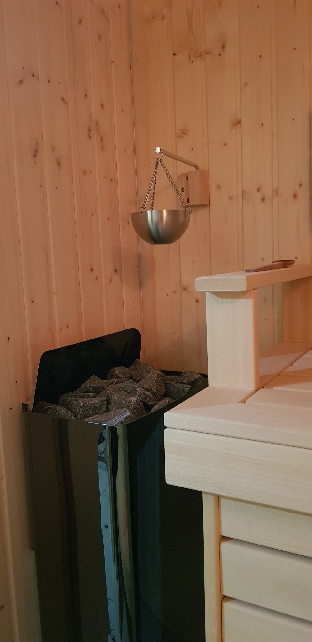 Wnętrze sauny z piecem wypełnionym kamieniami i drewnianymi ławkami. Na ścianie zawieszony metalowy czerpak na łańcuchu. Ściany wyłożone boazerią.