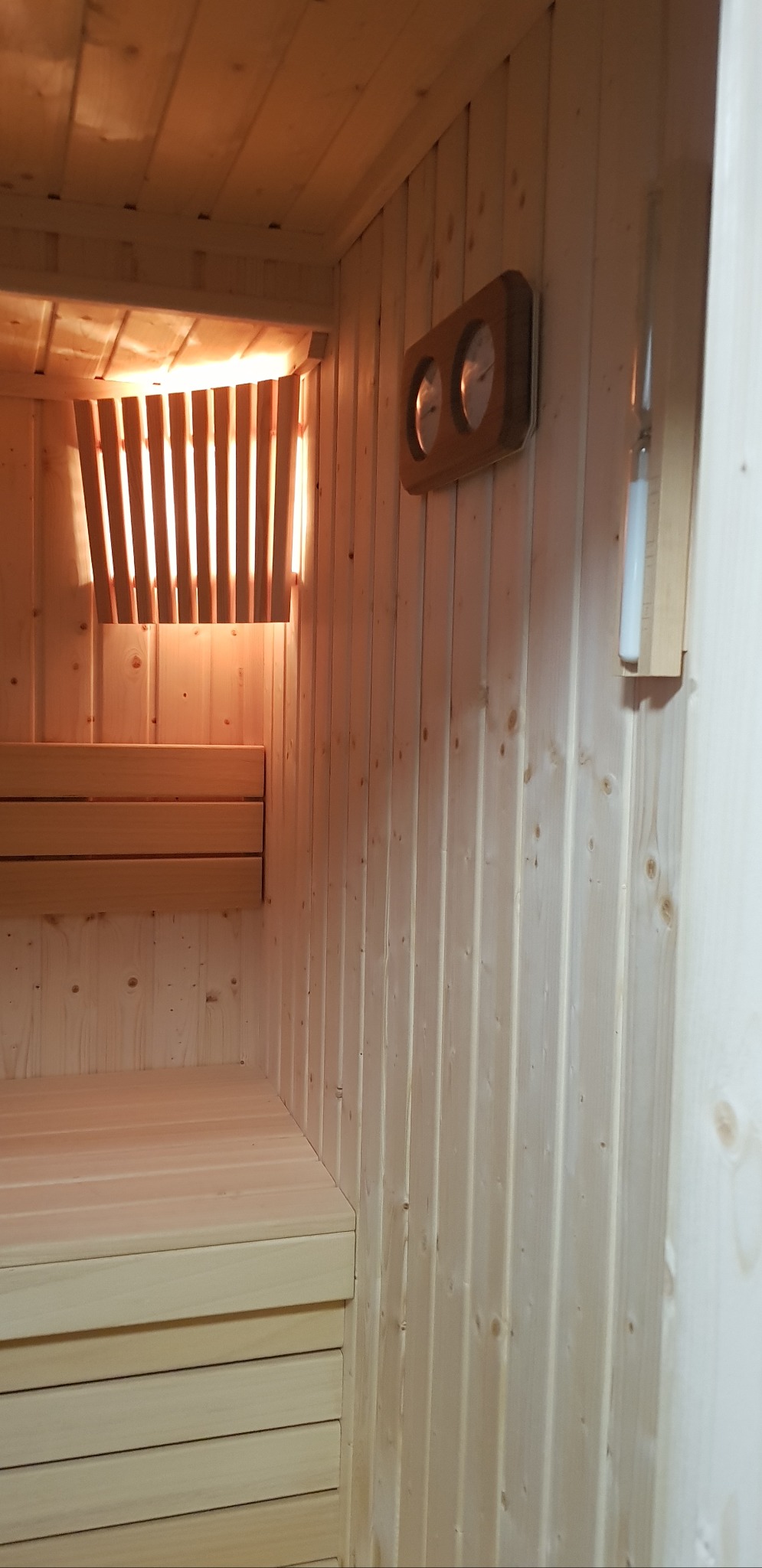 Jasne drewno w saunie: ławki, ściany z boazerii, osłona pieca z pionowych listew podświetlonych od tyłu, termometr i higrometr na ścianie.