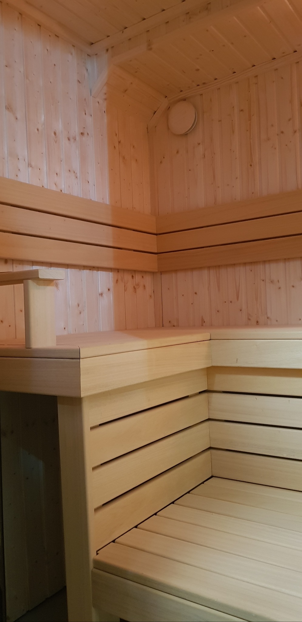 Jasne drewno w saunie: ławy z oparciem, boazeria ścienna i sufitowa. Okrągły, biały punkt świetlny na ścianie. Detal wykończenia.