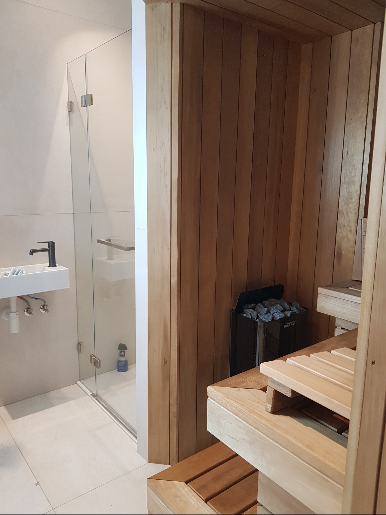 Nowoczesna sauna z drewnianym wykończeniem i szklaną kabiną prysznicową z czarną armaturą. Widoczny piec Harvia z kamieniami. Jasne, minimalistyczne wnętrze.