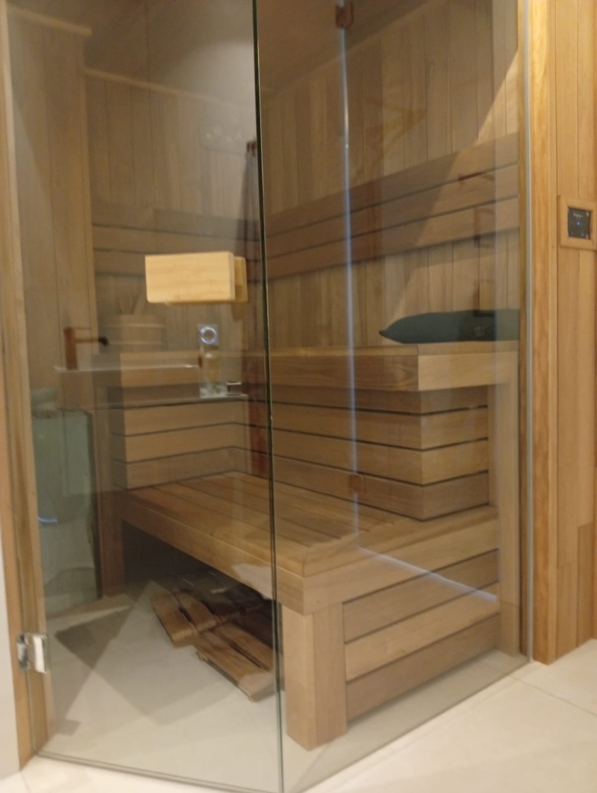 Szklana sauna z drewnianym wnętrzem. Widoczne ławki, oparcia i piec. Detale wykończenia z jasnego drewna. Nowoczesny design sauny.