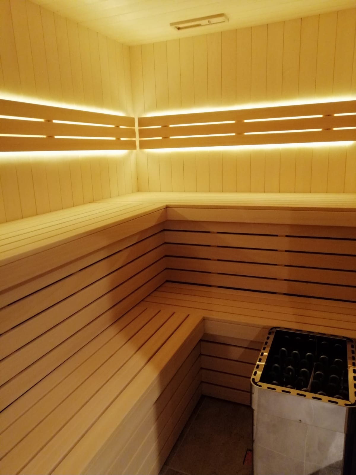 Jasne drewno i subtelne oświetlenie LED tworzą relaksującą atmosferę w saunie. Widoczne ławy i piec do sauny.