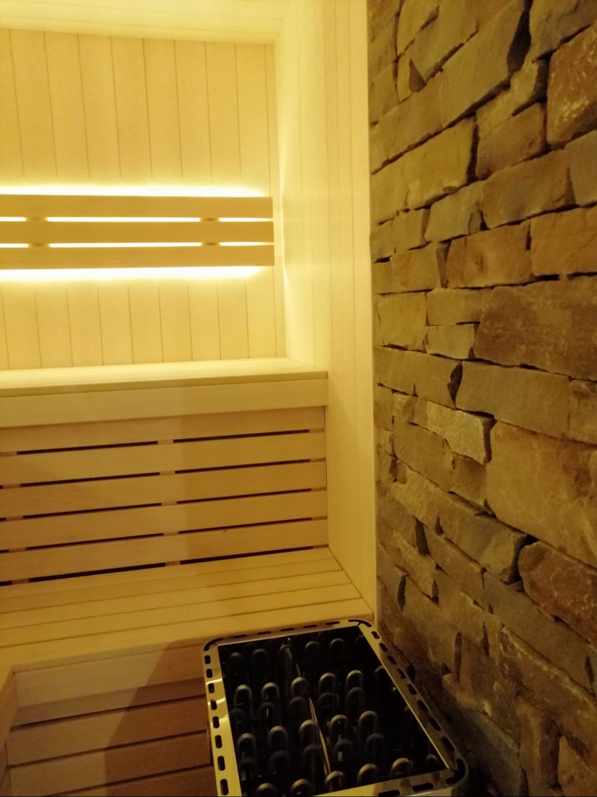 Jasne drewno sauny z podświetleniem LED i piecem, kontrastujące z kamienną ścianą. Widok z perspektywy wnętrza, ukazujący detale wykończenia i oświetlenia.