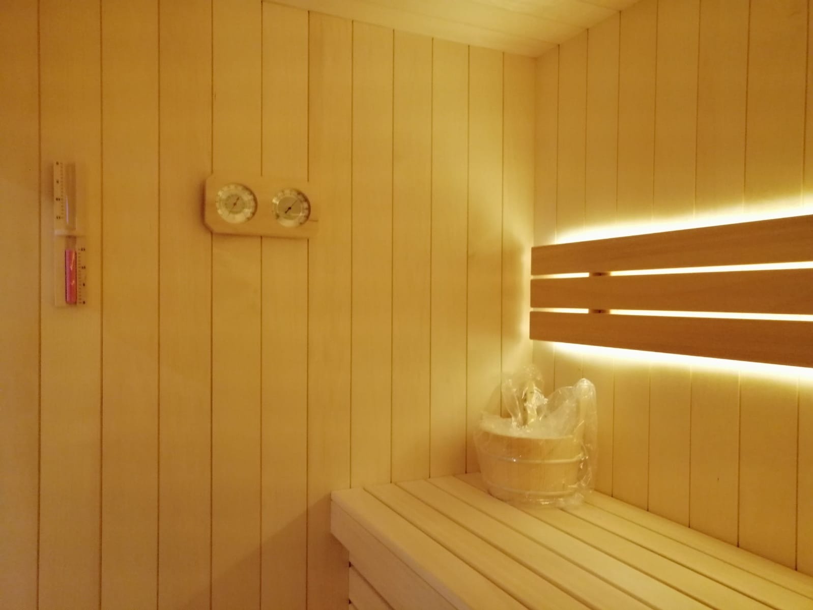 Jasne, drewniane wnętrze sauny z ławkami, termometrem i oświetleniem LED. Wiadro z wodą na ławce. Drewniane panele ścienne. Ciepłe, relaksujące światło.