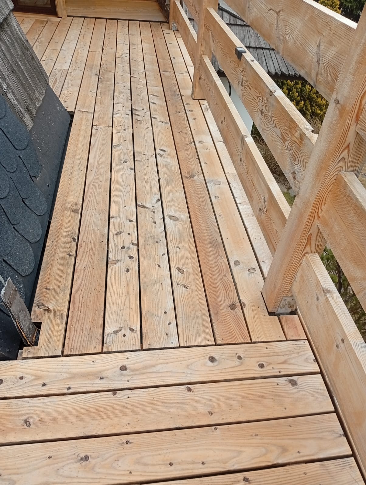 Jasny, drewniany taras z balustradą, widok z góry. Szare gonty dachu i zieleń w tle. Naturalne drewno z widocznymi słojami. Solidna konstrukcja tarasu.