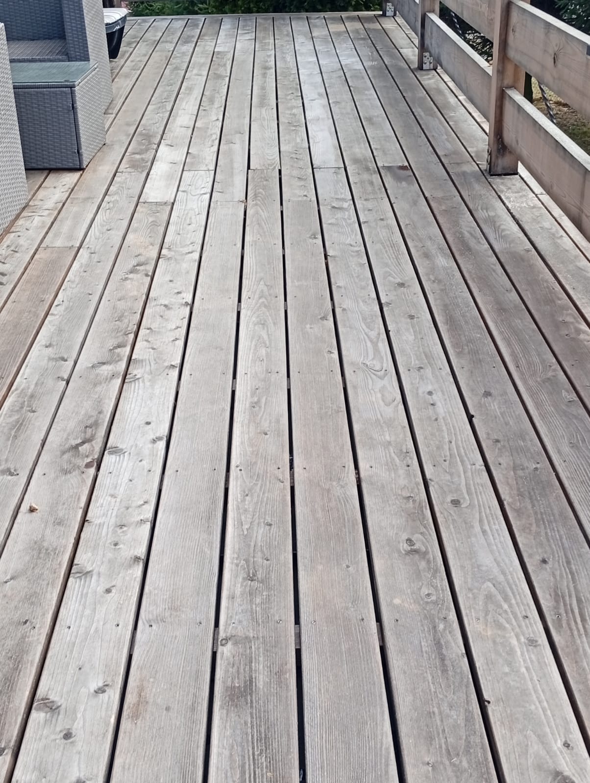 Szary taras drewniany z widoczną fakturą desek i fragmentem balustrady. W tle zieleń. Perspektywa z góry.