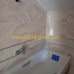 ReVa Anatolii Reva - Wanna obudowana płytkami z dekoracyjnym pasem, widoczna bateria i otwór w ścianie z wystającymi przewodami.