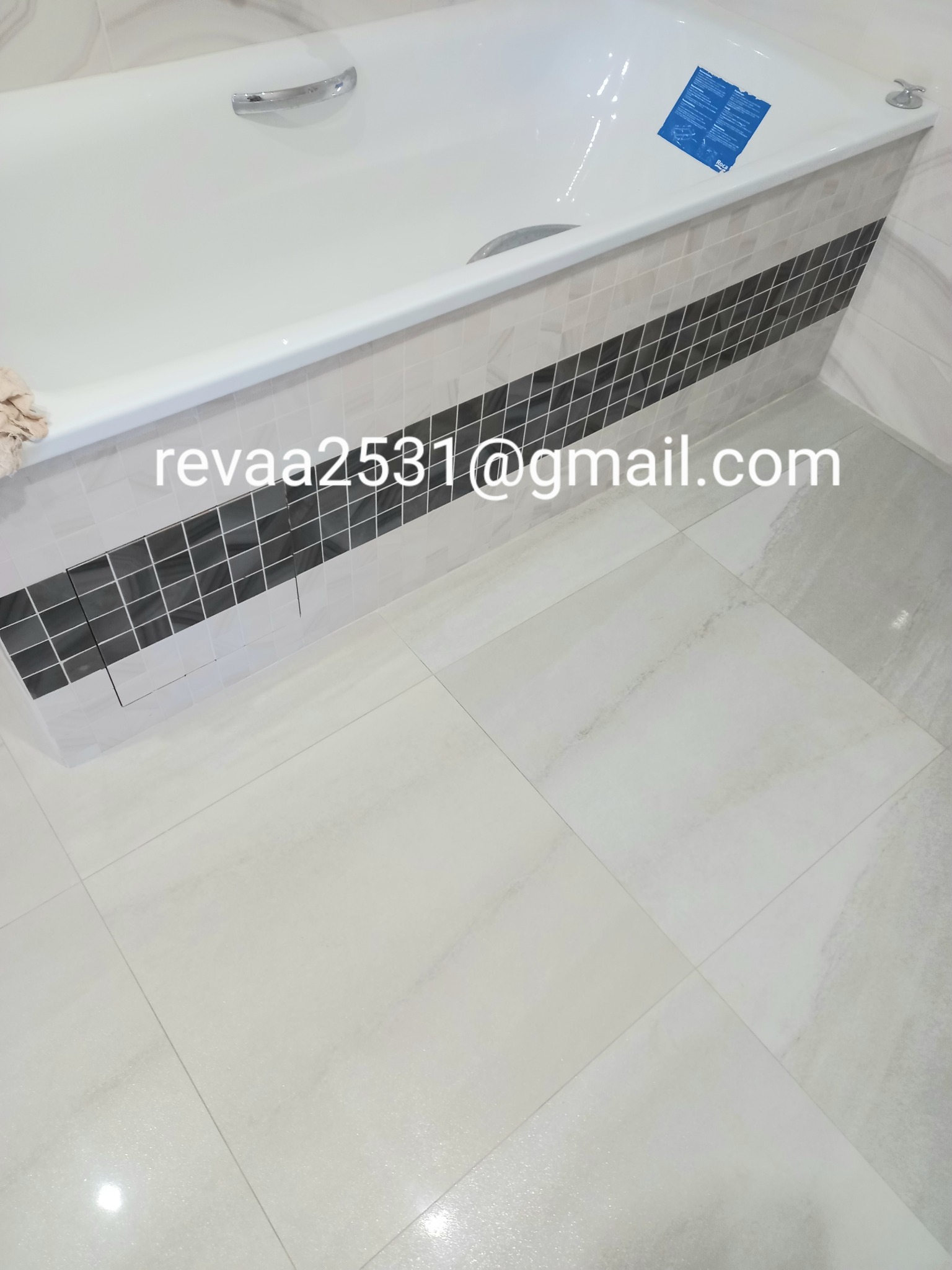 Nowa, biała wanna z chromowaną armaturą, obłożona jasnymi płytkami z dekoracyjnym pasem czarnych mozaik, oraz podłoga wyłożona jasnymi, marmurkowymi płytkami.