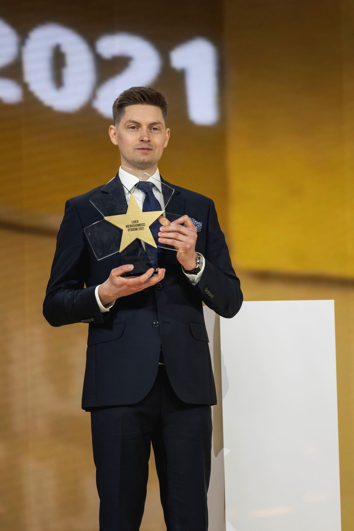 Mężczyzna w garniturze trzymający nagrodę w kształcie gwiazdy z napisem 'Lider Nieruchomości Otodom 2021' na tle sceny z napisem '2021'.