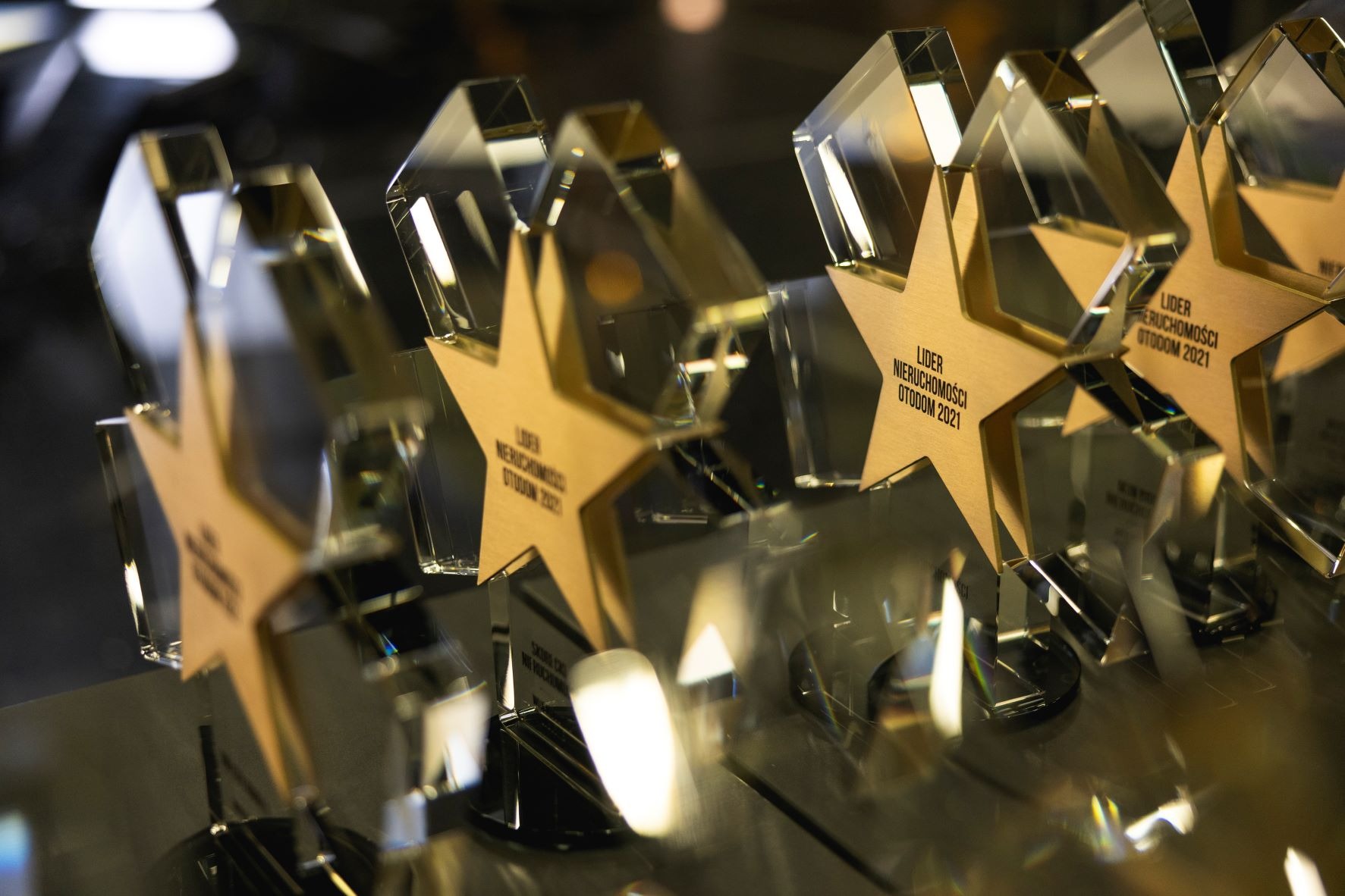 Szereg szklanych statuetek w kształcie gwiazdy z napisem 'Lider Nieruchomości Otodom 2021' na złotym tle, ustawionych na ciemnej, błyszczącej powierzchni.