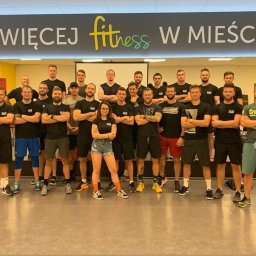 Marcin Kubica - trener personalny, trener przygotowania motorycznego - Grupowe zdjęcie instruktorów fitness w czarnych koszulkach z logo, stojących pod banerem z napisem 'Największej Fitness w Mieście', w tle widoczny sprzęt do ćwiczeń.