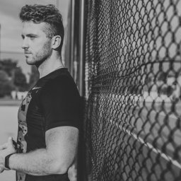 Marcin Kubica - trener personalny, trener przygotowania motorycznego - Mężczyzna w sportowej koszulce opiera się o metalową siatkę na zewnątrz, z zamyślonym wyrazem twarzy i splecionymi dłońmi, w tle widoczny fragment boiska.