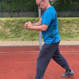Marcin Kubica - trener personalny, trener przygotowania motorycznego - Mężczyzna w okularach, w niebieskiej koszulce i szarych spodniach dresowych, wykonuje ćwiczenia rozciągające dłonie na czerwonej bieżni lekkoatletycznej.