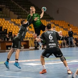 Marcin Kubica - trener personalny, trener przygotowania motorycznego - Mężczyzna w zielonej koszulce z numerem 27 skacze w górę z piłką do piłki ręcznej, otoczony przez obrońców w czarno-zielonych strojach na tle żółtych trybun hali sportowej.