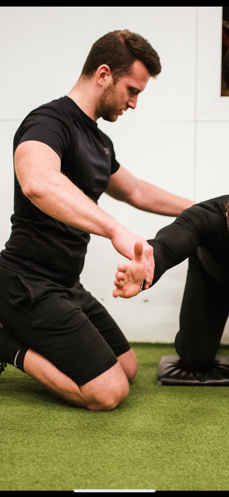 Mężczyzna w czarnym stroju sportowym, klęczący na zielonej nawierzchni, pomaga drugiej osobie w rozciąganiu ręki.