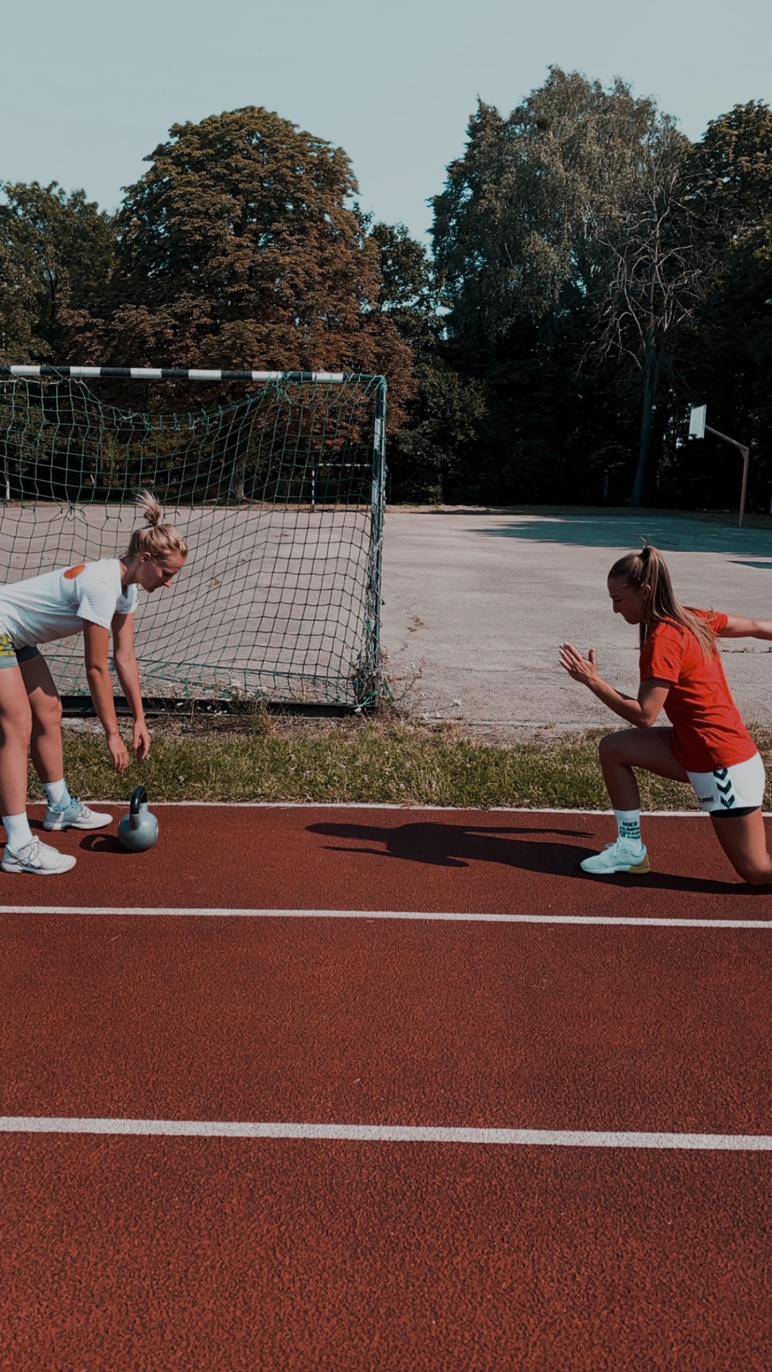 Dwie kobiety ćwiczą na bieżni, jedna z kettlebellem, druga w pozycji startowej, w tle bramka do piłki nożnej i drzewa.