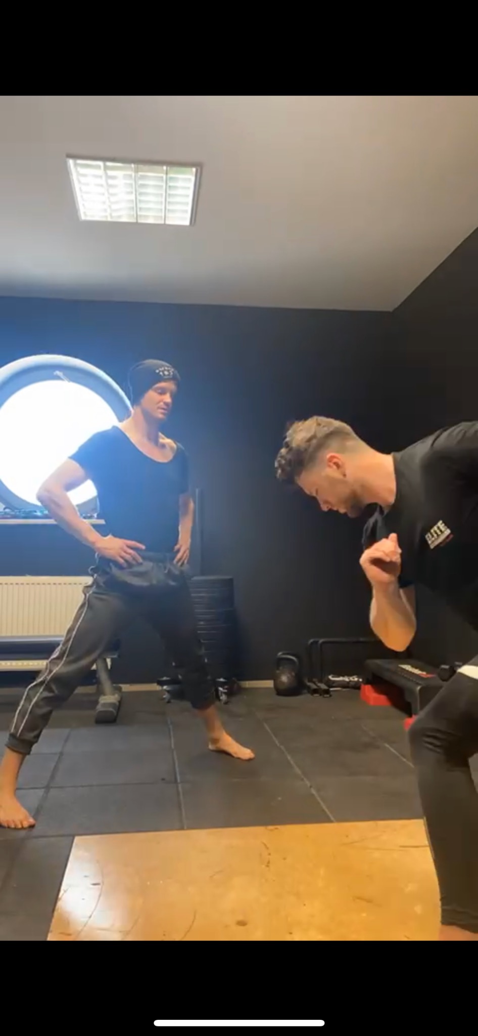 Mężczyzna w czarnej koszulce i spodniach dresowych, z opaską na głowie, wykonuje rozciągające ćwiczenie nóg w siłowni, podczas gdy drugi mężczyzna w czarnym stroju sportowym obserwuje, z widocznym...