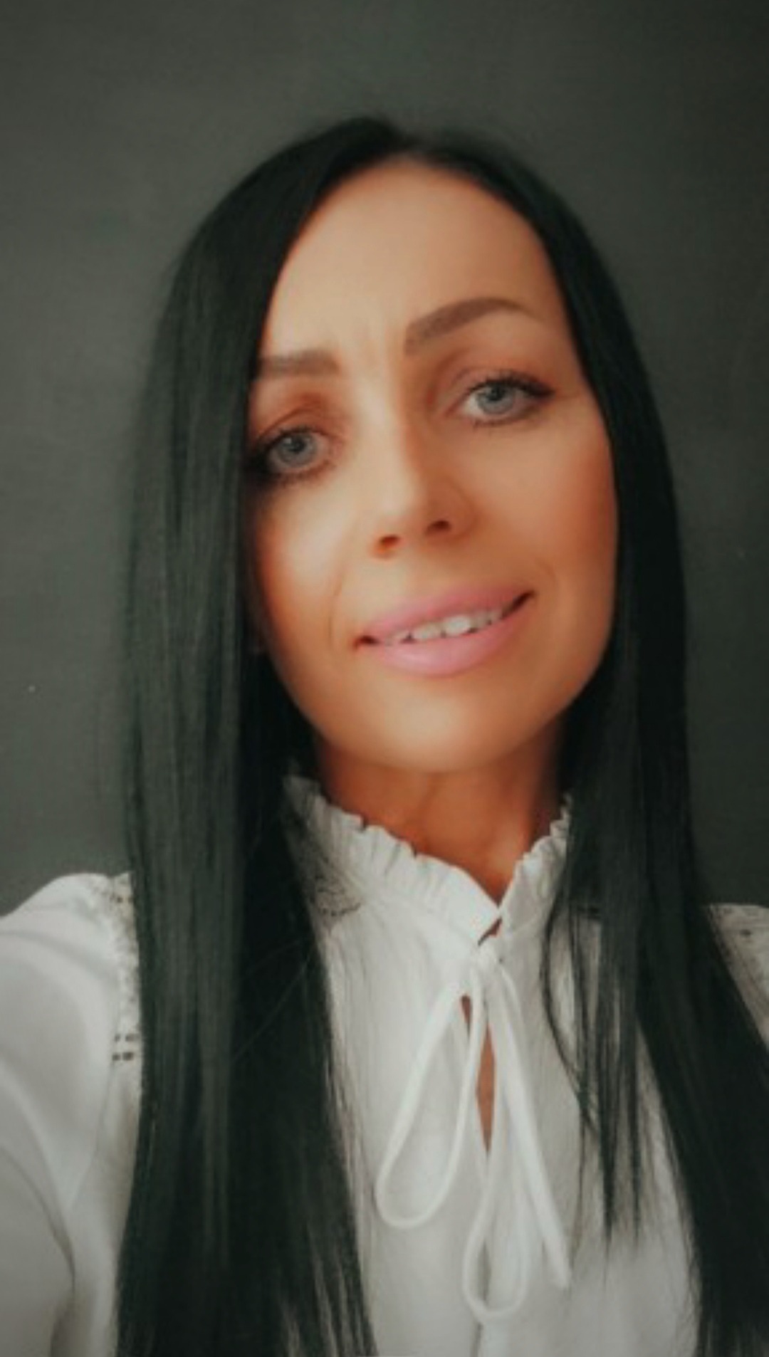 Portret uśmiechniętej kobiety z długimi, czarnymi włosami i jasnymi oczami, ubrana w białą bluzkę z wiązaniem pod szyją, na ciemnoszarym tle.