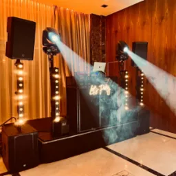 Profesjonalny sprzęt nagłośnieniowy i oświetleniowy z laptopem na stanowisku DJ-a, z neonowym napisem 'Let's Party', oświetlony reflektorami i otoczony efektem mgły na parkiecie.