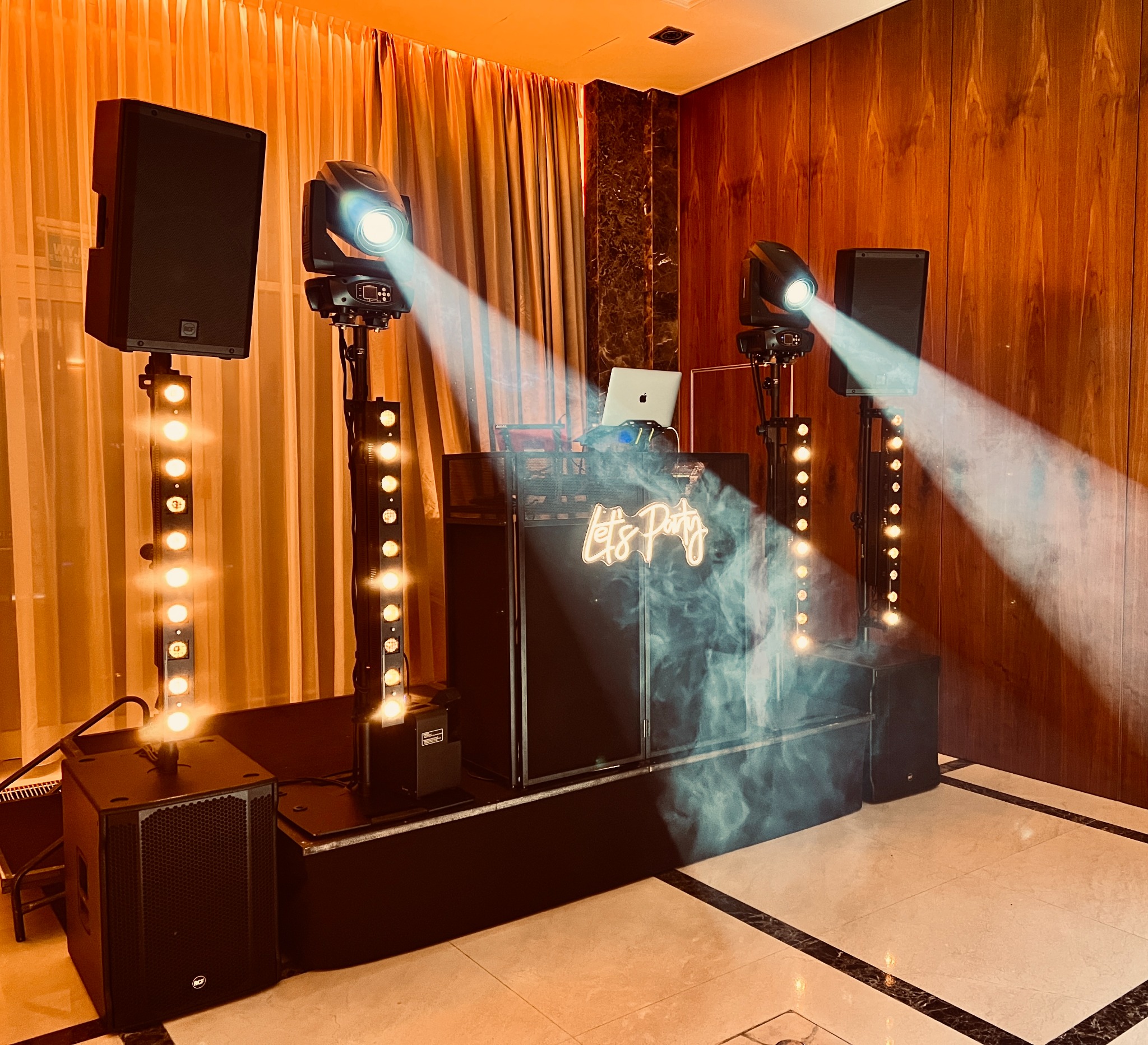 Profesjonalny sprzęt nagłośnieniowy i oświetleniowy z laptopem na stanowisku DJ-a, z neonowym napisem 'Let's Party', oświetlony reflektorami i otoczony efektem mgły na parkiecie.