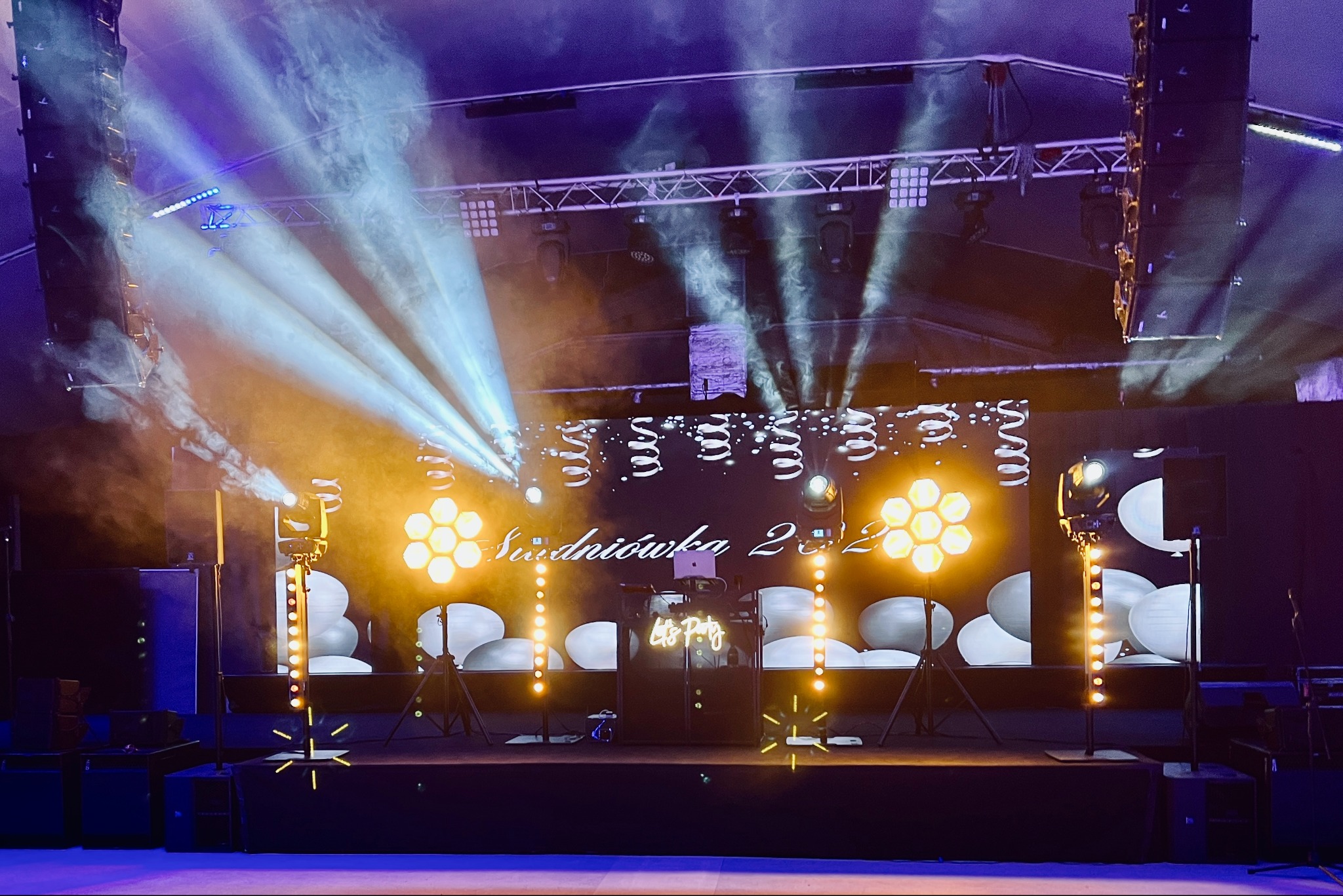 Scena z nagłośnieniem i oświetleniem podczas imprezy, widoczne kolumny głośnikowe, reflektory, dekoracje balonowe i napis 'Studniówka 2022' na ekranie, dym w tle.