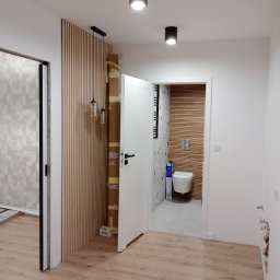 Ivan Pidhirnyi LW-STROJ - Wnętrze mieszkania w trakcie remontu: widok na wykończoną łazienkę z drewnianą okładziną, panele ścienne w przedpokoju i kartony z materiałami budowlanymi przy ścianie.