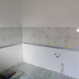 Ivan Pidhirnyi LW-STROJ - Ściana w trakcie układania płytek z użyciem listwy montażowej i krzyżyków dystansowych. Widoczne wystające kable i rury instalacyjne.