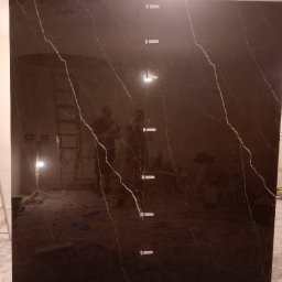 Ivan Pidhirnyi LW-STROJ - Duża płyta z ciemnego marmuru z białymi żyłami, ustawiona pionowo, z widocznymi elementami mocującymi i odbiciem sylwetek osób w tle.