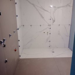 Ivan Pidhirnyi LW-STROJ - Wnętrze łazienki w trakcie remontu: białe płytki imitujące marmur na ścianach, z niebieskimi krzyżykami dystansowymi, szare ściany boczne i podłoga w trakcie prac.