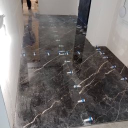 Ivan Pidhirnyi LW-STROJ - Układanie ciemnych, marmurkowych płytek podłogowych z użyciem poziomujących klipsów i klinów wzdłuż fug.