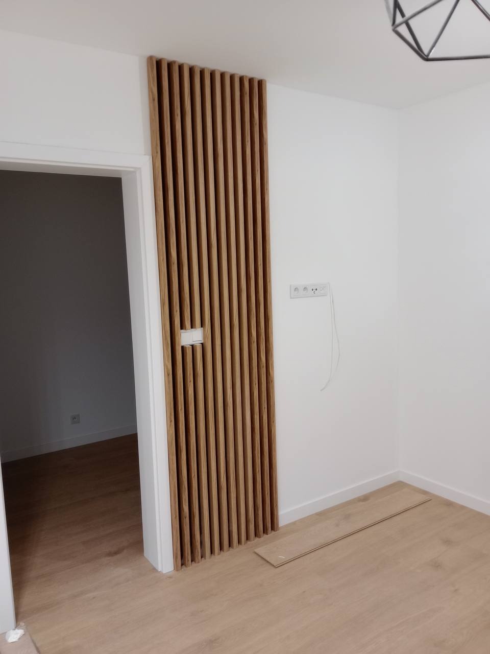 Nowoczesny pokój z drewnianą ścianką działową z pionowych listew, jasną podłogą i białymi ścianami, widoczne gniazdka elektryczne i fragment niezakończonej podłogi.